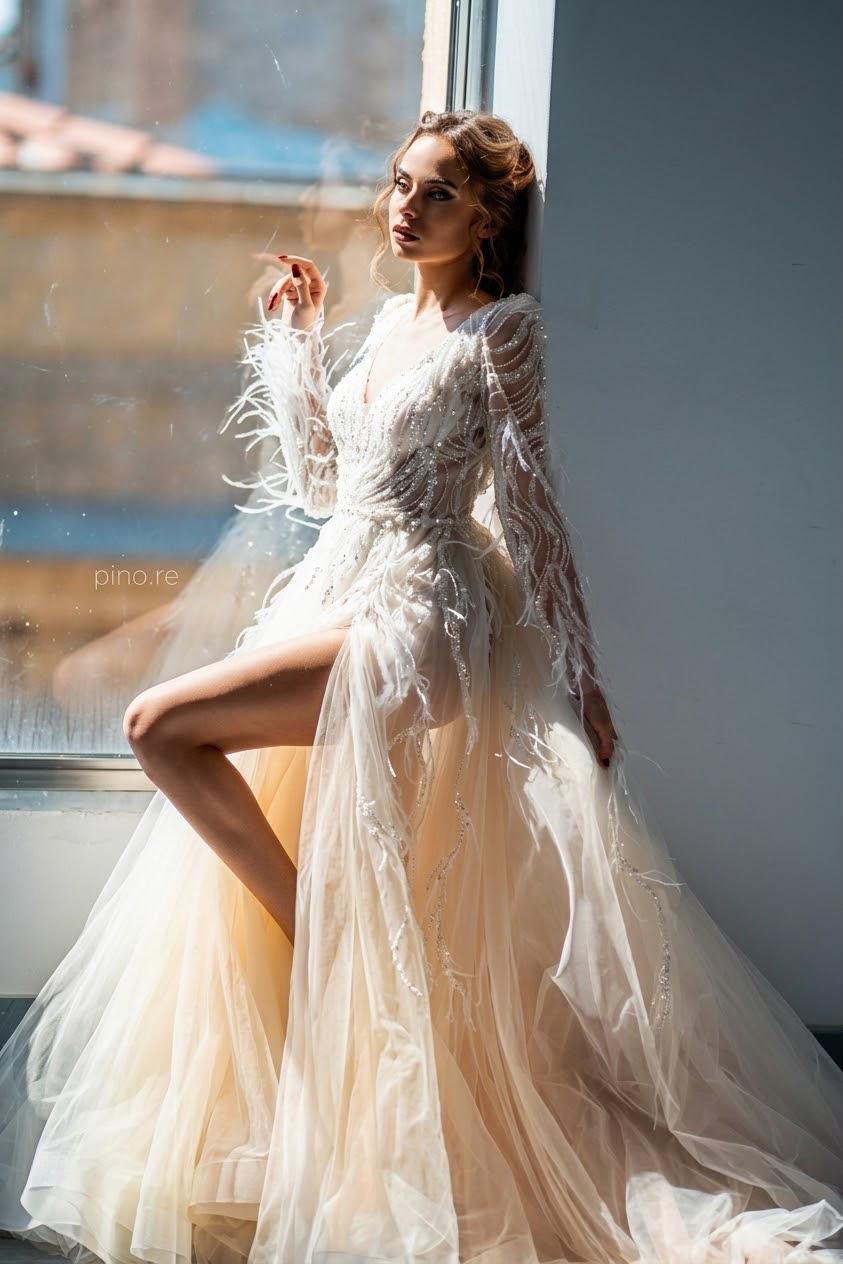 Sposa in abito di tulle con piume e ricami accanto alla finestra